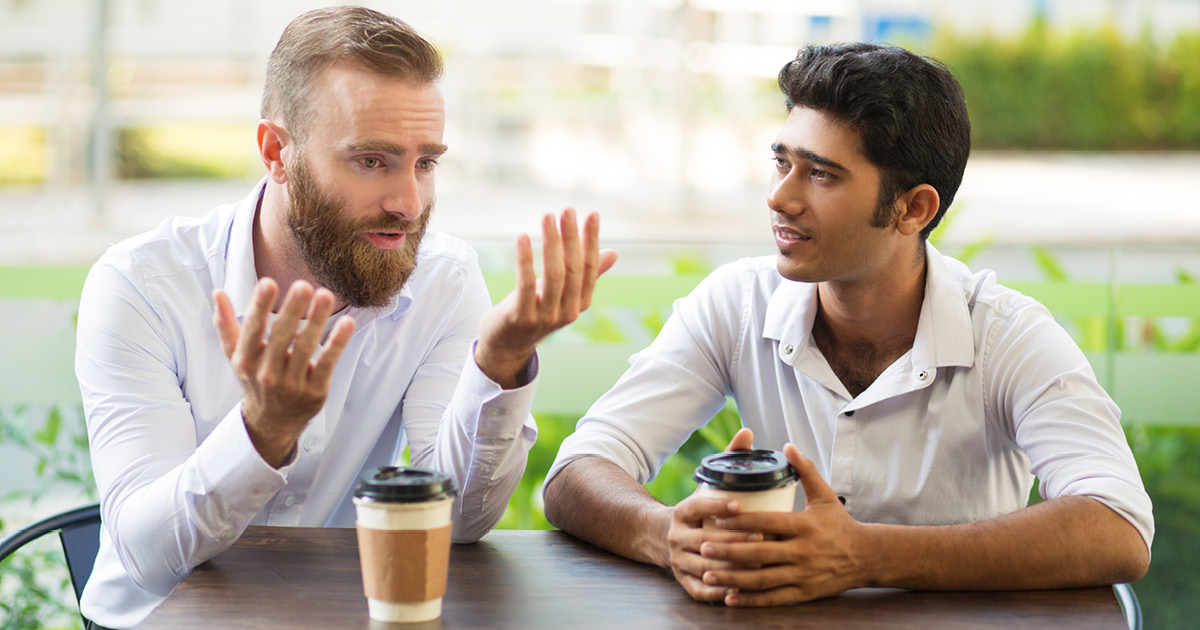 Two people sit at a table outdoors, engaging in conversation. Both have coffee cups. The person on the left gestures expressively as the other listens intently.