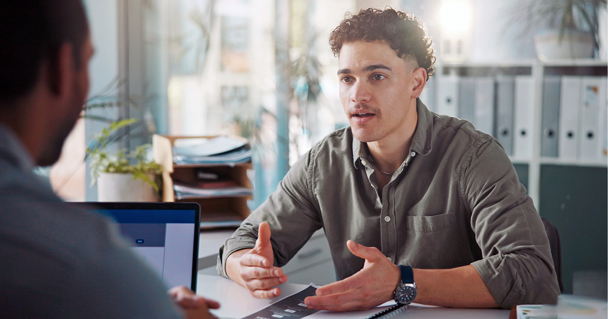 Two people talking to each other as they sit on opposite sides of a desk in an office.